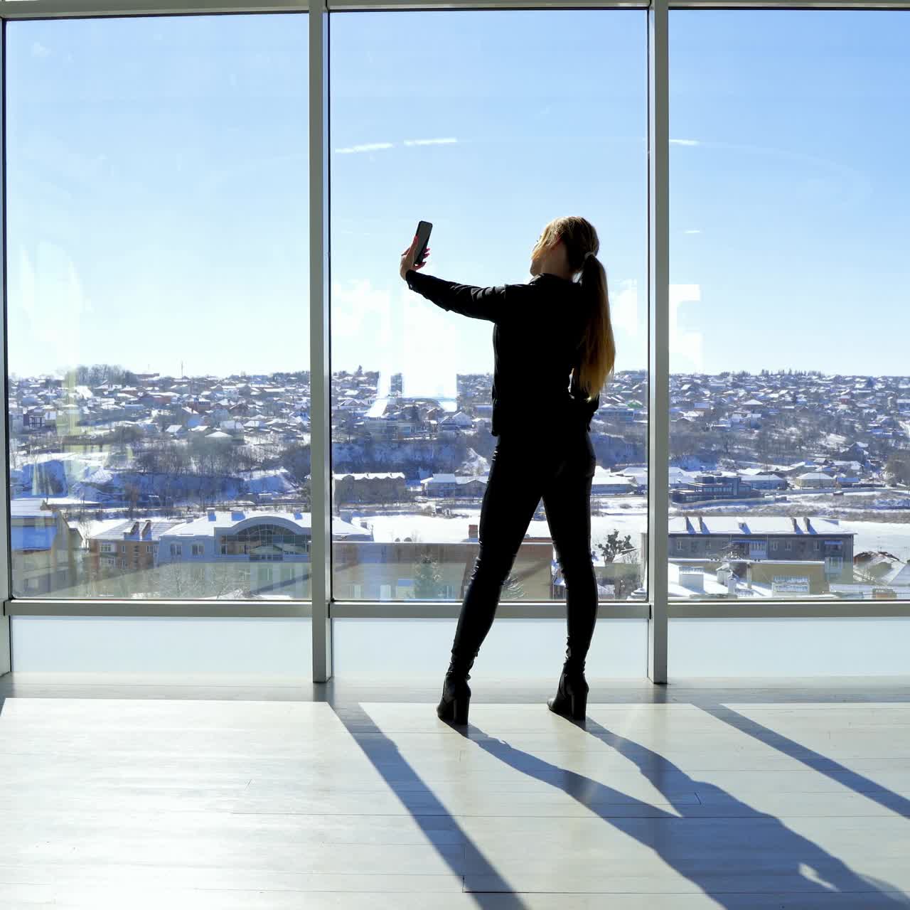 Girl is doing selfie in office. Young woman making photos of herself on a mobile phone while standing near the large office window in a sunny day. Rear view