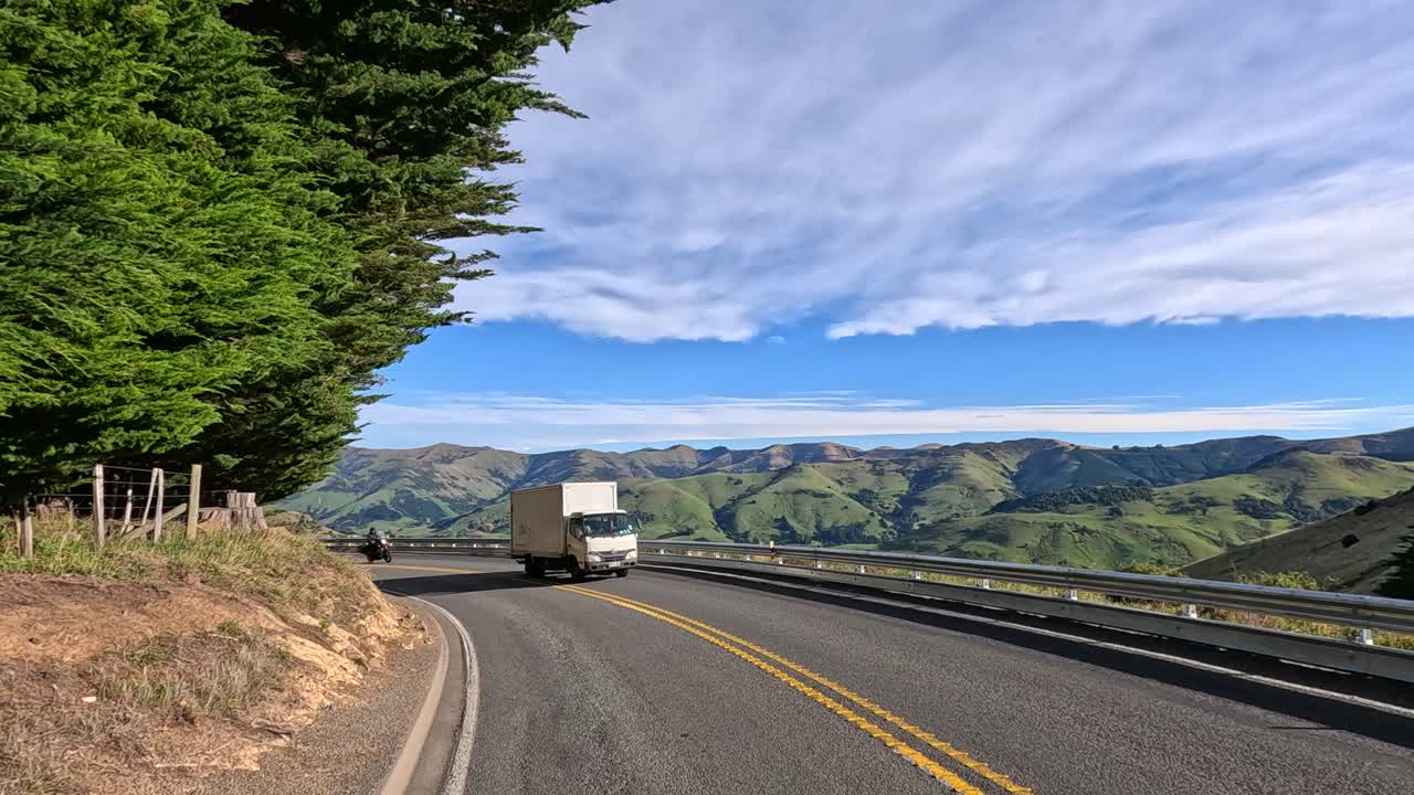 Vehicles traverse a winding road in Akaroa, New Zealand, under clear skies and lush green hills, capturing serene rural landscapes