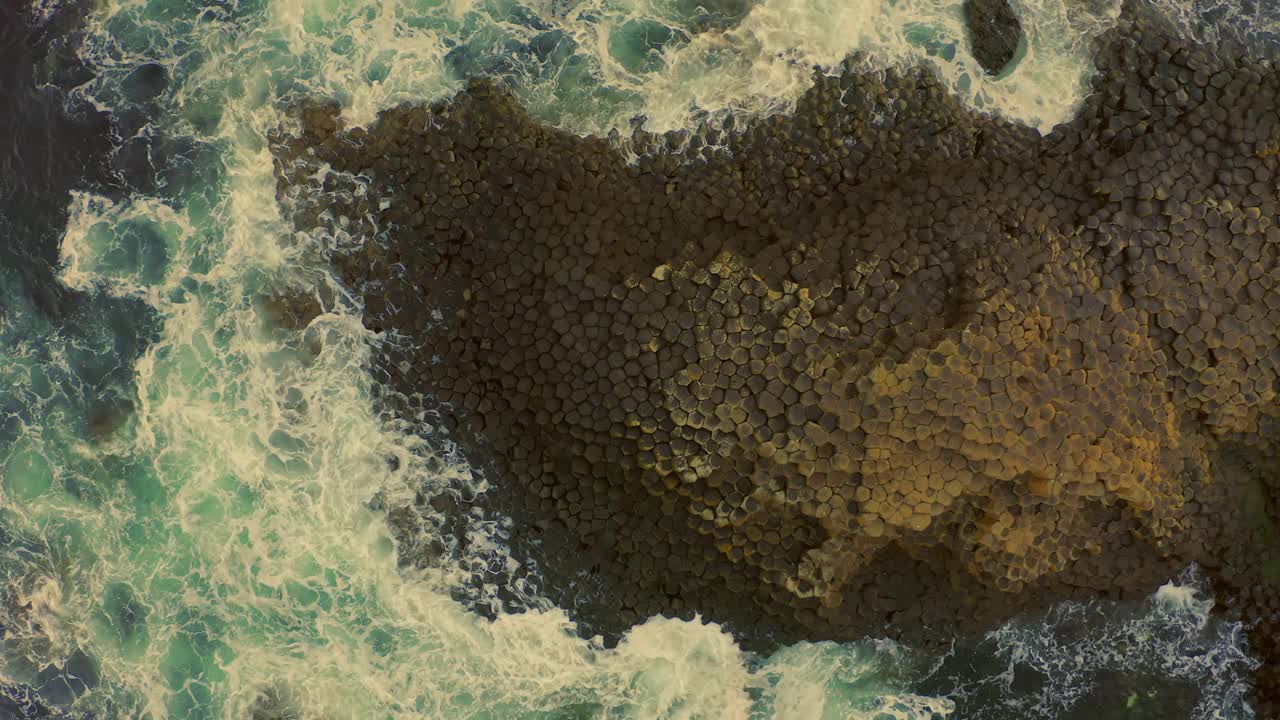Drone descends over the famous honeycomb rock formation at Giants Causeway, waves crashing as the shot moves from wide to close-up