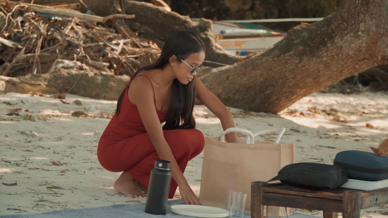 Coppia che condivide una cerimonia del tè in spiaggia, donna in abito rosso prepara le tazze da un cesto intrecciato mentre il compagno con un cappello casual osserva la riva sabbiosa, legno trasportato dalla corrente, bicchieri, thermos isolante, cuscini, atmosfera calda.