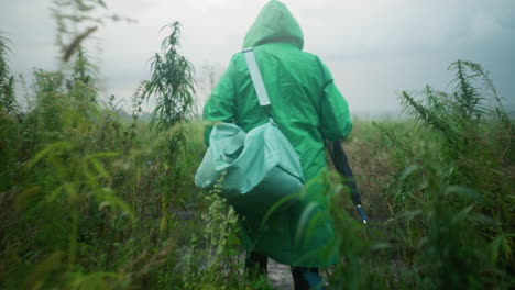 back view of individual in green raincoat carrying mint-colored bag and umbrella, wearing yellow rain boots, walking carefully along a path surrounded by tall greenery and plants