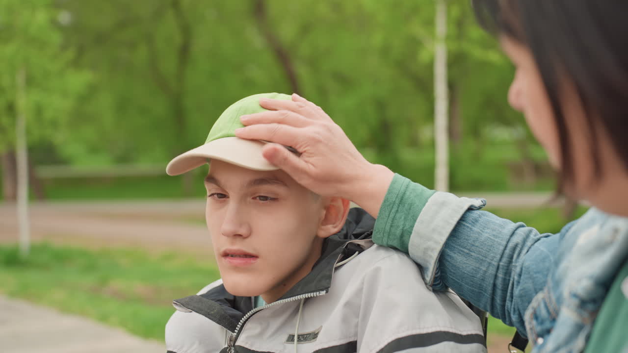 Gentle Caregiver Touch, Woman Cares For Young Teen, Compassionate Lady Assists Boy On Wheelchair, Kind Woman Gently Adjusts Hat On Young Boy, Caring Individual Lovingly Helps Teen With Hat Outside