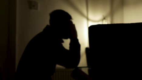 silhouette of distressed young man sat at table with atmospheric lighting, with his head in hands