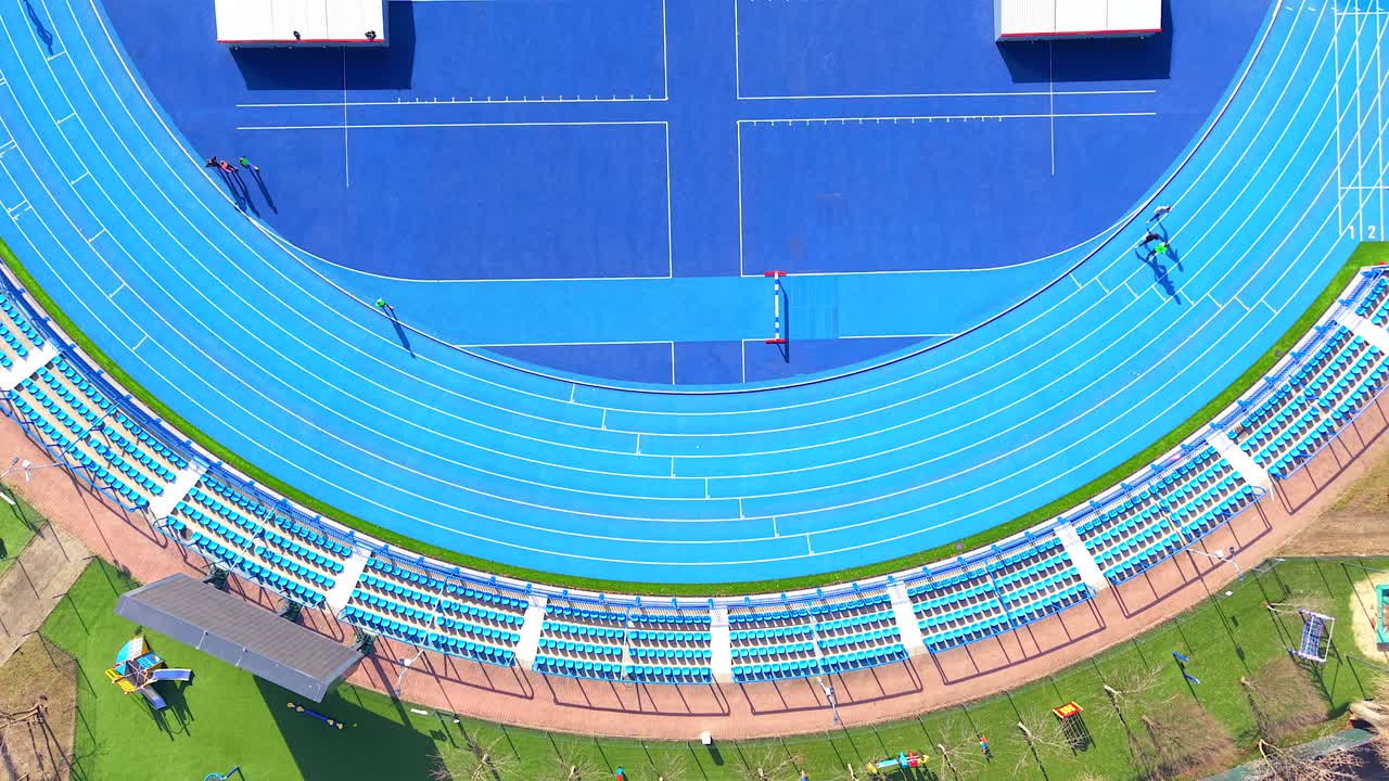 Luftfoto ovenfra av idrettsutøvere som løper på en moderne blå løpebane på et idrettsstadion. Det livlige idrettsfeltet er omgitt av tomme seter.