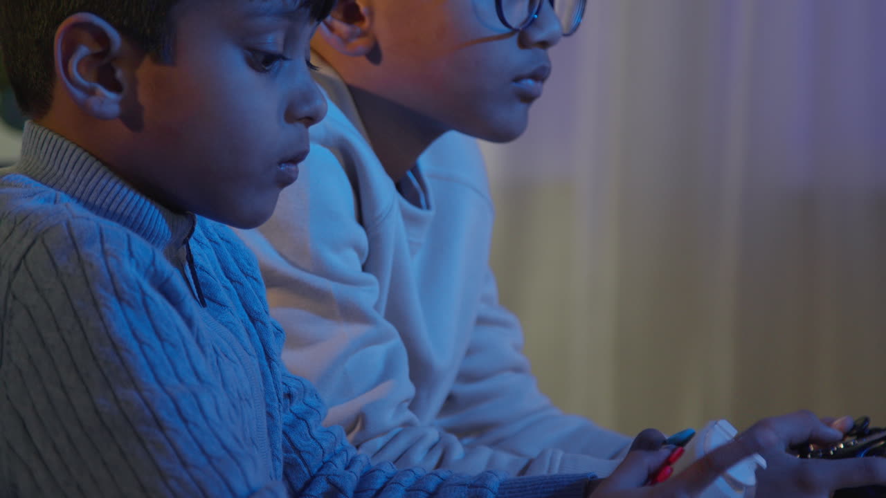 Close Up Of Two Young Boys At Home Playing With Computer Games Console On TV Holding Controllers Late At Night