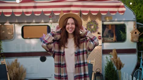 Portrait-of-a-happy-brunette-girl-in-a-brown-hat-in-a-pink-plaid-shirt-who-adjusts-her-clothes-posing-and-smiling-Near-her-trailer-outside-the-city-during-her-picnic-at-the-camp-in-the-summer