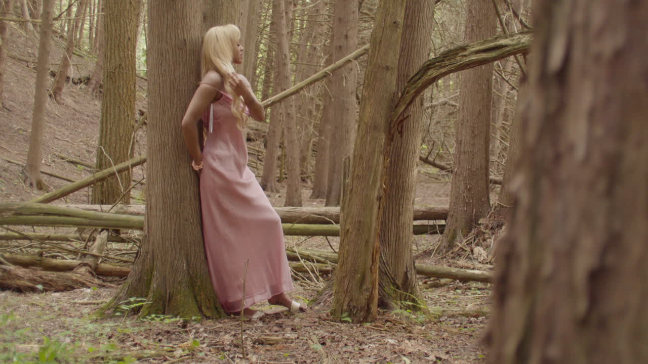 joven mujer de ébano, con el cabello largo rubio, con un vestido rosa. se apoya contra el tronco de un árbol de goma de azúcar, en un bosque. juega con su cabello como ella mira a, y lejos de la cámara, tiro amplio.