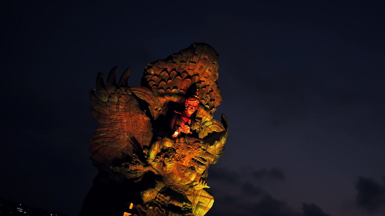 illuminated monument rising high above Bali island skyline at red dusk sunset showcasing giant statue scale and dramatic travel landmark architecture aerial drone view