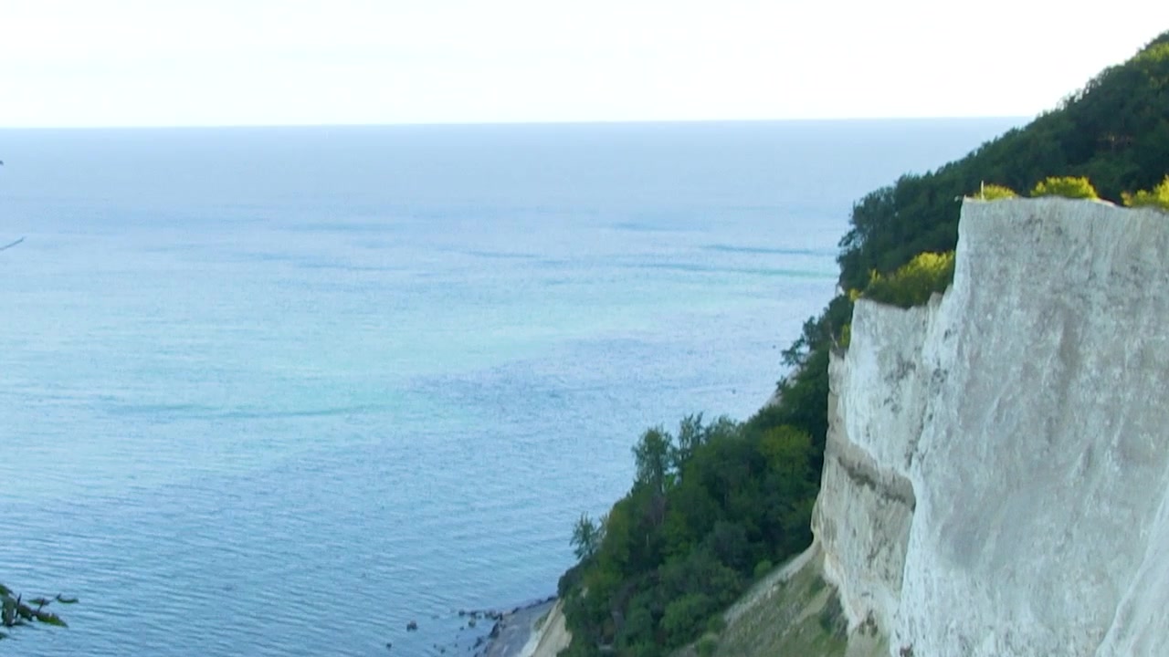 Tilting up from where the white cliffs of Mons Klint in Denmark meet the blue and green ocean, up to a view of the open sea.