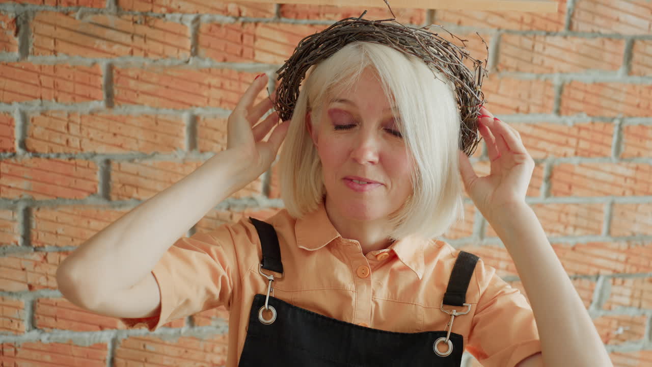 Female florist with blonde hair in apron smiling while holding handcrafted twig wreath in front of face against rustic brick wall, showcasing creativity and joy of floral decorative craft work indoors