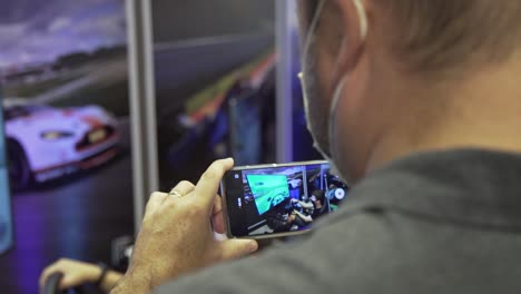 over the shoulder of a man holding a phone filming a guy playing in a racing simulator cockpit