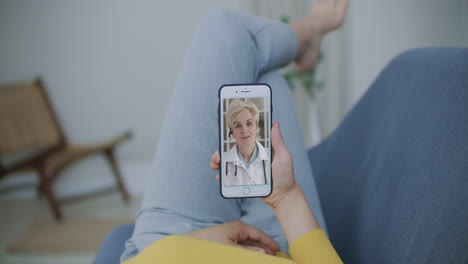 A woman lying on the couch talking to a woman doctor on video using a smartphone. Remote medical consultation with a doctor via mobile phone