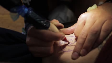 foto de cerca de un artista del tatuaje mientras apuñala hábilmente las líneas de un tatuaje con tinta negra usando una máquina de tatuaje con aguja y mirando la plantilla en la piel
