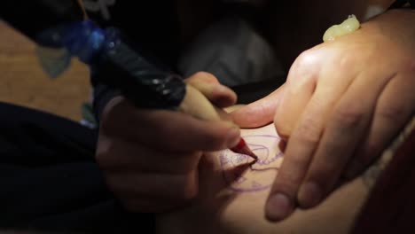 handheld close up shot of a tattoo artist as he skillfully stabs the lines of a tattoo with black ink using a tattoo machine with needle and looking at the stencil on the skin