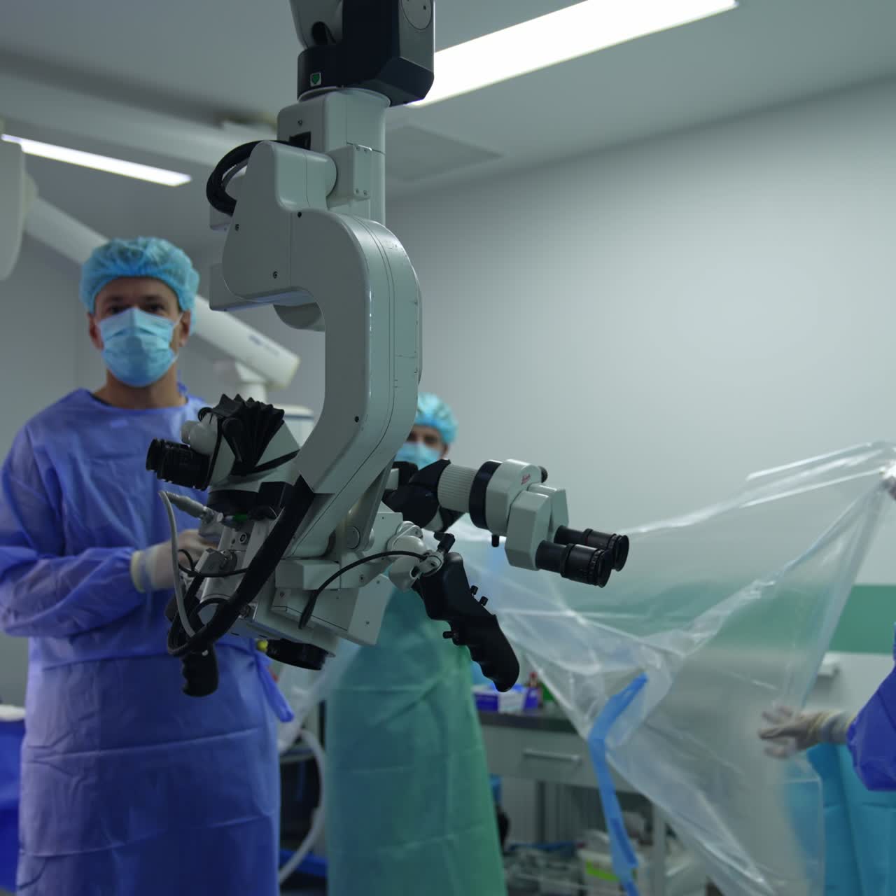 Mid-aged male surgeon turns away from operational table and speaks to the camera. Medical assistants spread the plastic cover for the equipment
