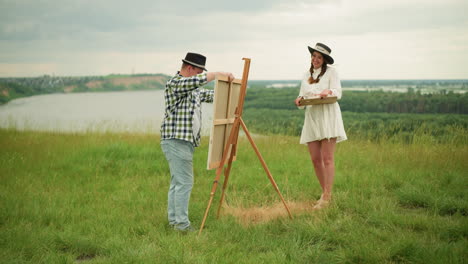 un artista con un sombrero negro y una camisa a cuadros está montando una tabla de dibujo en un trípode de madera en un campo cubierto de hierba. una mujer con un vestido blanco está cerca, sonriendo mientras observa la configuración