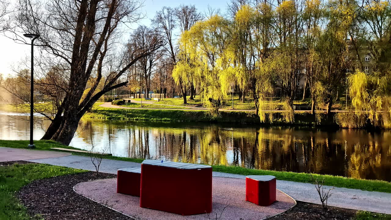 Park benches and trees reflected in still water of city canal in Valmiera, Latvia