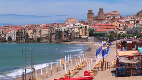 a small boat and umbrellas rest on a beach with houses near the shoreline in cefalu italy