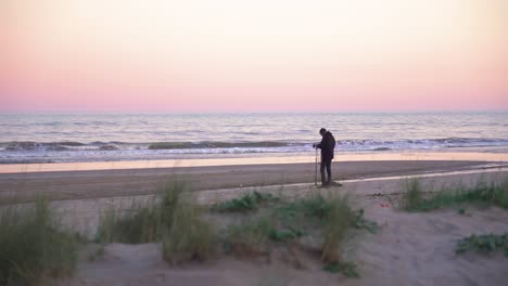 Sound-technician-is-recording-environments-on-the-beach-at-sunset,-the-pink-sky-and-the-waves-of-the-sea-as-a-motive-with-the-horizon-in-the-background-of-the-sequence