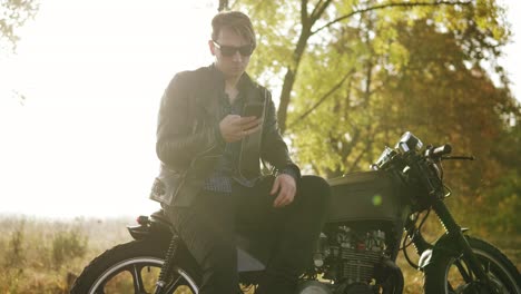 Young-attractive-man-in-leather-jacket-and-stylish-sunglasses-is-standing-by-his-bike-on-the-country-road-on-a-sunny-day-in-autumn-against-the-sun-and-using-his-phone