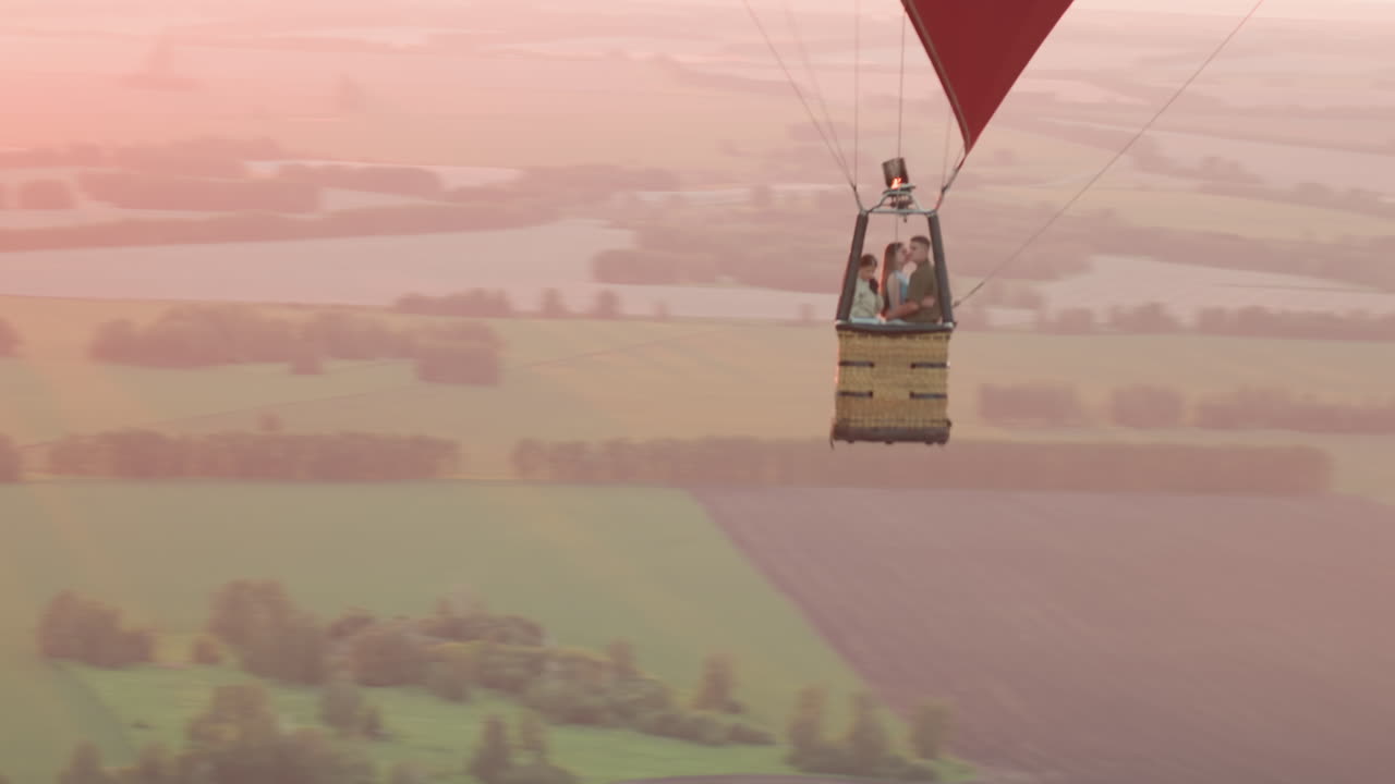 travelers in colorful hot air balloon drifting over expansive green farmland at sunset while pilot ignites burner flame adjusting altitude capturing serene sky adventure from aerial perspective