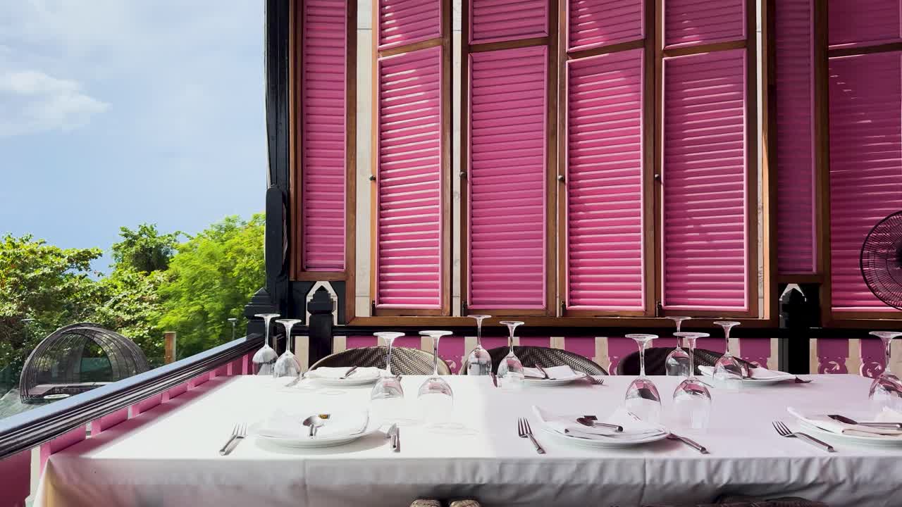 Brightly colored restaurant with pink shutters and set tables, overlooking lush greenery in Phuket, Thailand. Daytime, vibrant atmosphere