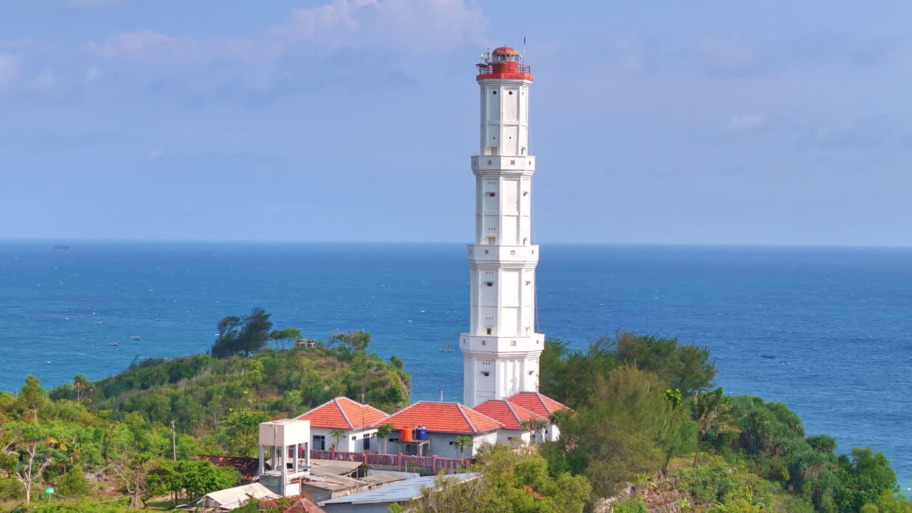 Scenic drone shot of a coastal lighthouse surrounded by tropical vegetation, facing the vast horizon of the ocean. Baron Beach Lighthouse, Indonesia