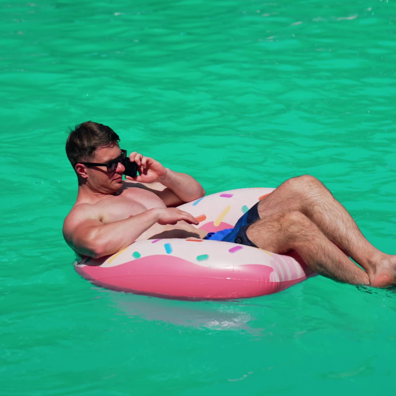 Sunbathing Caucasian man sitting in the inflatable circle talking on the phone. Male wearing sunglasses resting outdoors
