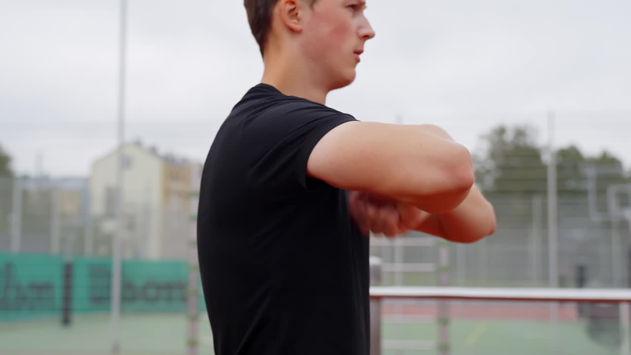 Young man stretching arms and shoulders outdoors on a sports court, preparing for exercise, fitness training, or warm-up before athletic activity