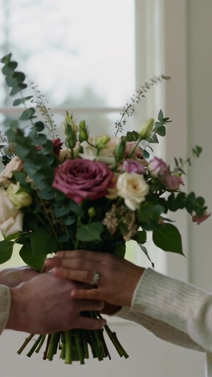 Video verticale: Una mano entra in scena, una persona con una camicia chiara sistema e porge un bouquet al proprio partner a casa.