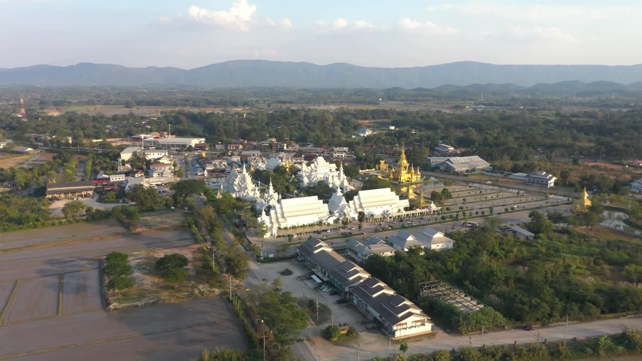 drone aéreo al revés de wat rong khun el enorme templo budista blanco y templo dorado con montañas y paisaje en chiang rai, tailandia