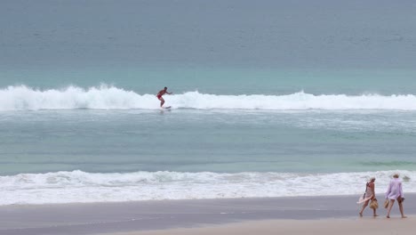 a surfer catches waves as people walk on the beach.