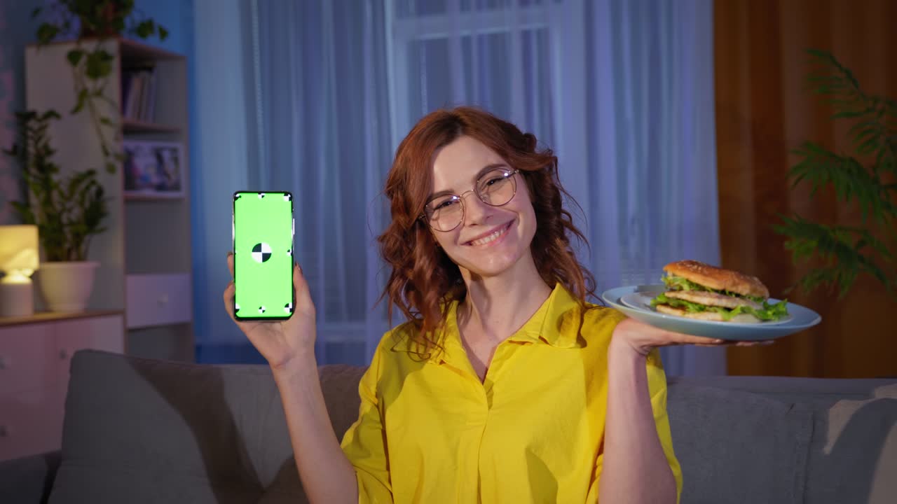 fast food delivery, portrait of charming smiling woman with burger and smartphone with green chroma key on screen sits on sofa in room in evening smiling and looking at camera