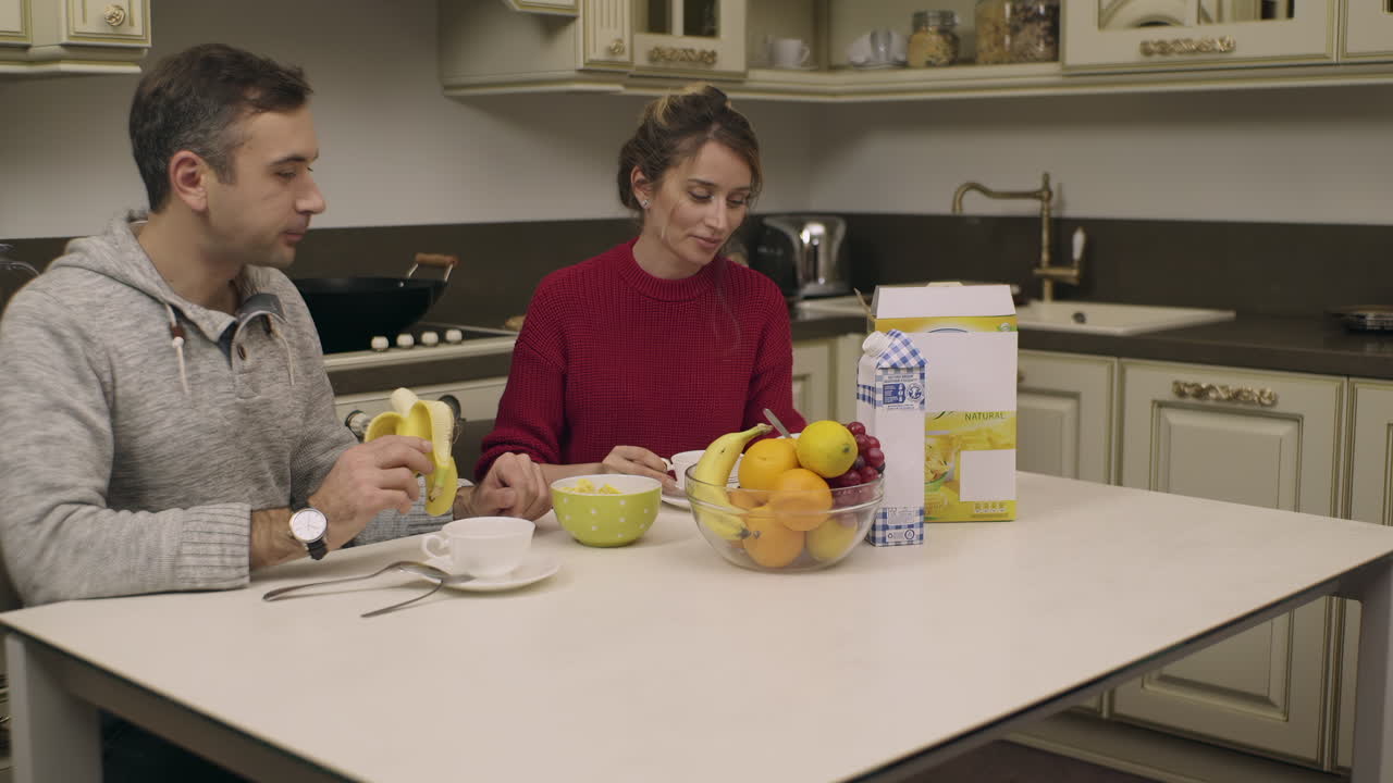 A man and woman sit at a kitchen table enjoying breakfast. They share fruits and tea while engaging in friendly conversation and laughter. The atmosphere is warm and inviting