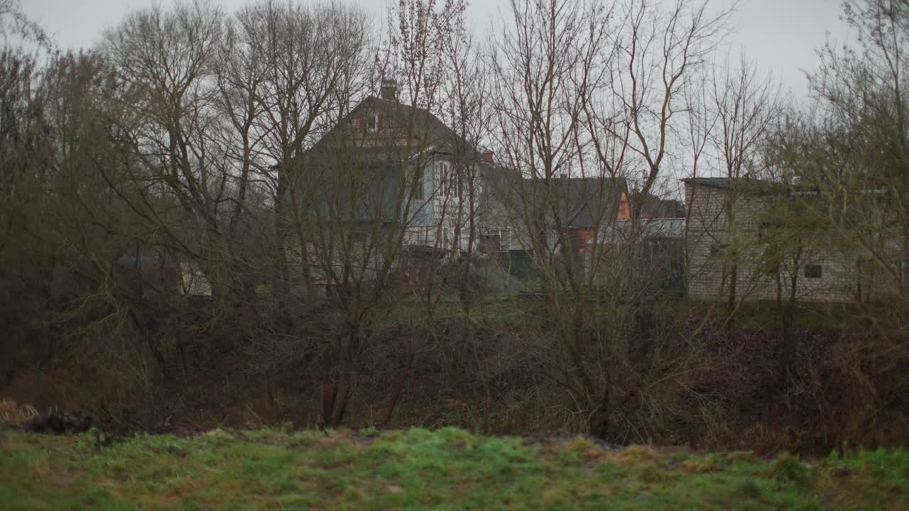 Casa rurale in legno ed edifici annessi in mattoni visti attraverso gli alberi spogli invernali sotto un cielo nuvoloso. Tipica architettura dei villaggi dell'Europa orientale in inverno