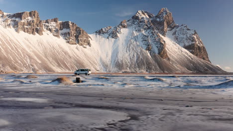 toma de seguimiento de vuelo de drones de una furgoneta blanca que llega a la enorme montaña vestrahorn en islandia durante el día soleado - rocas cubiertas de nieve y paisaje durante el frío invierno