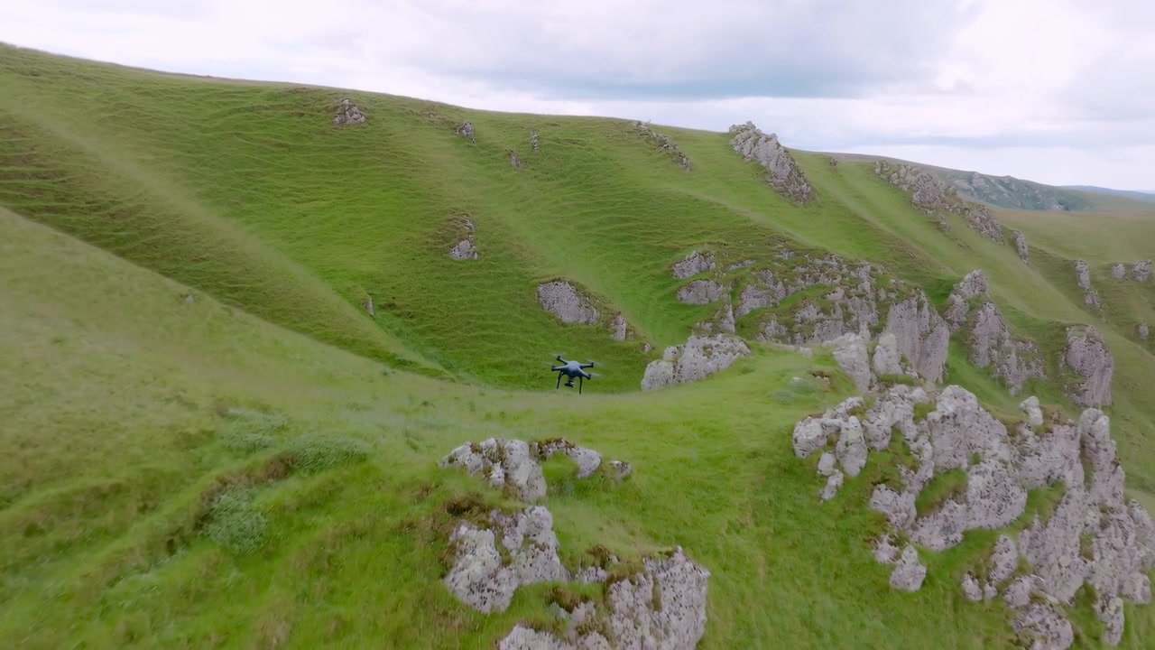 Un piccolo drone si muove attraverso un vasto paesaggio rurale sopra erba luminosa, formazioni rocciose chiare e pendii ampi, creando un'atmosfera di viaggio all'aperto calma con un'estetica commerciale pulita e un ampio scenario.