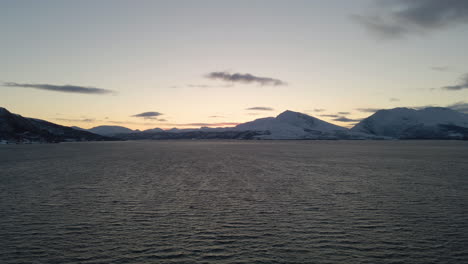 Arctic-drone-flight-over-ocean-at-blue-hour,-golden-glow-behind-snowy-mountains