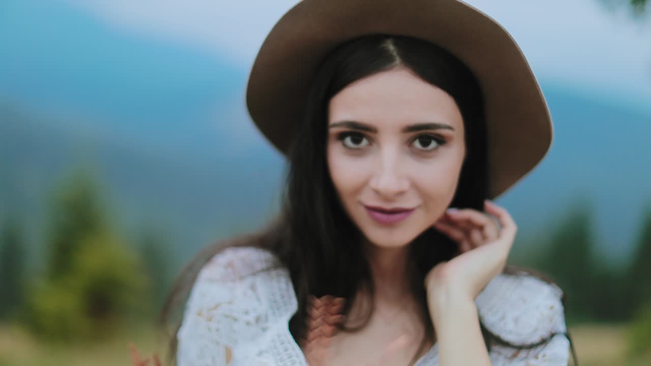 Portrait of brunette in hat among nature. Attractive young woman looking at camera and smiling on blur nature background.