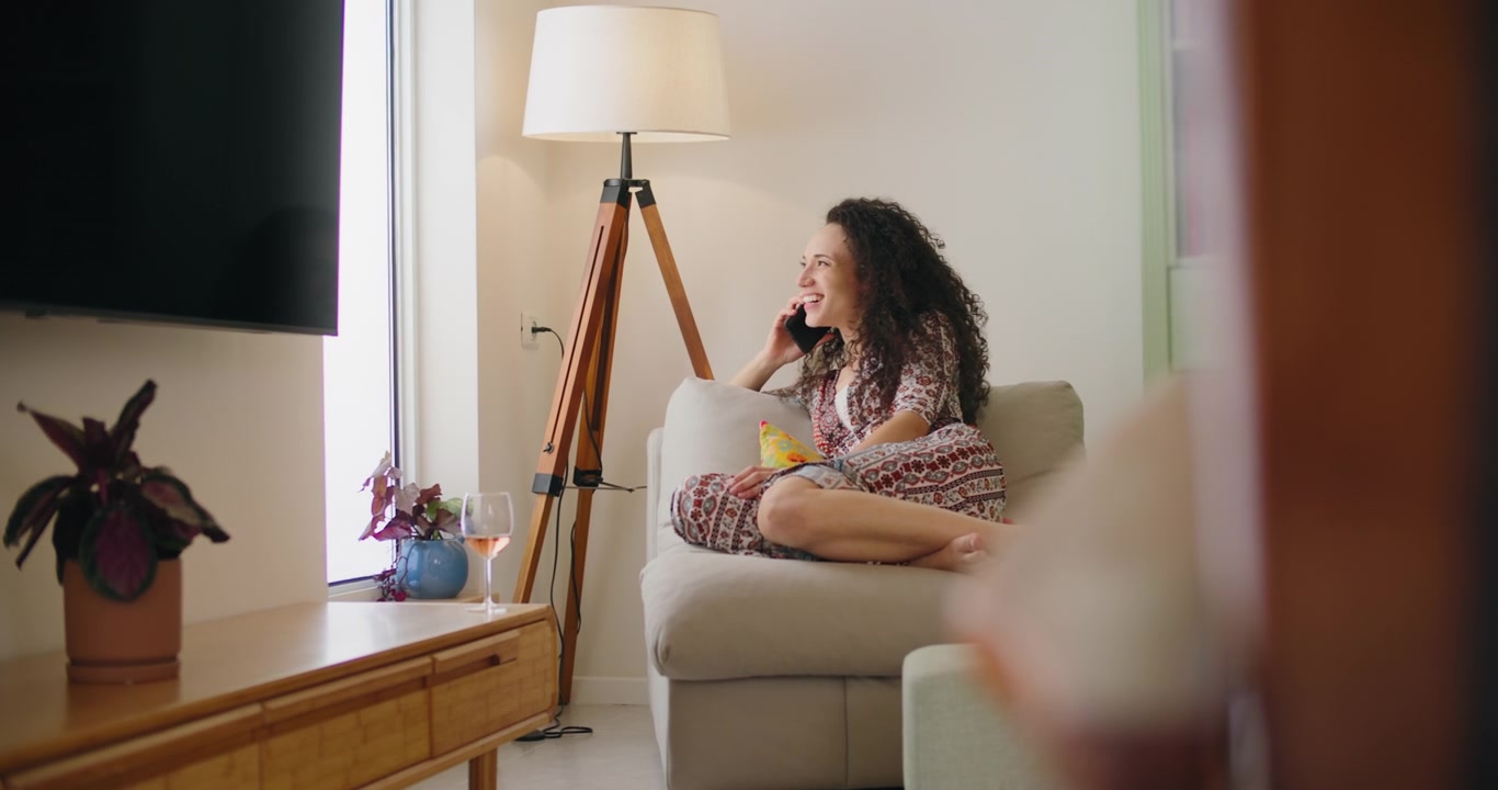 Ung kvinne slapper av på sofaen og snakker i smarttelefon hjemme
