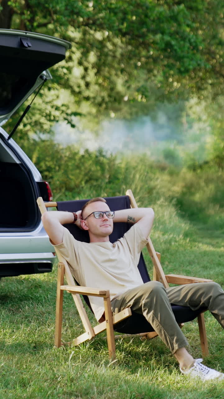 Man sitting comfortably in a folding chair stretches hands up. Summer relaxation in the wood. Vertical video