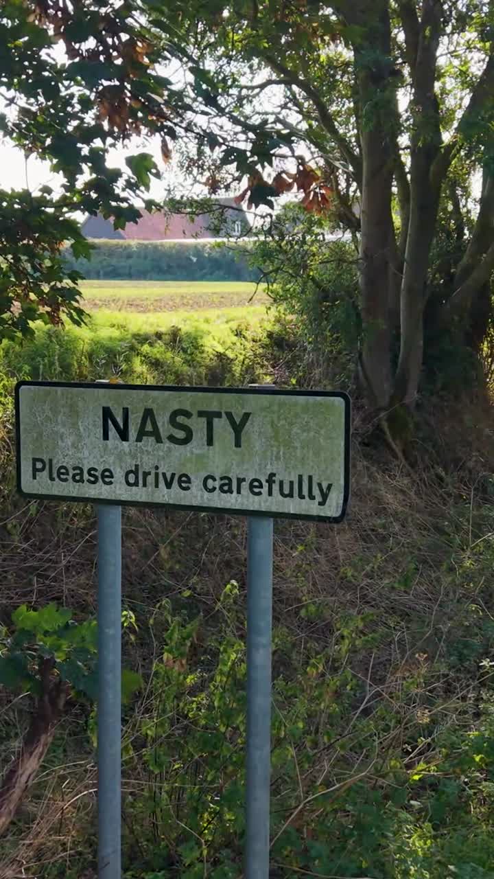 Vertical camera tracks right across the Nasty, Hertfordshire hamlet sign, ending side on. Shafts of light filter through surrounding trees, creating a blend of rural charm, atmosphere, and metaphor