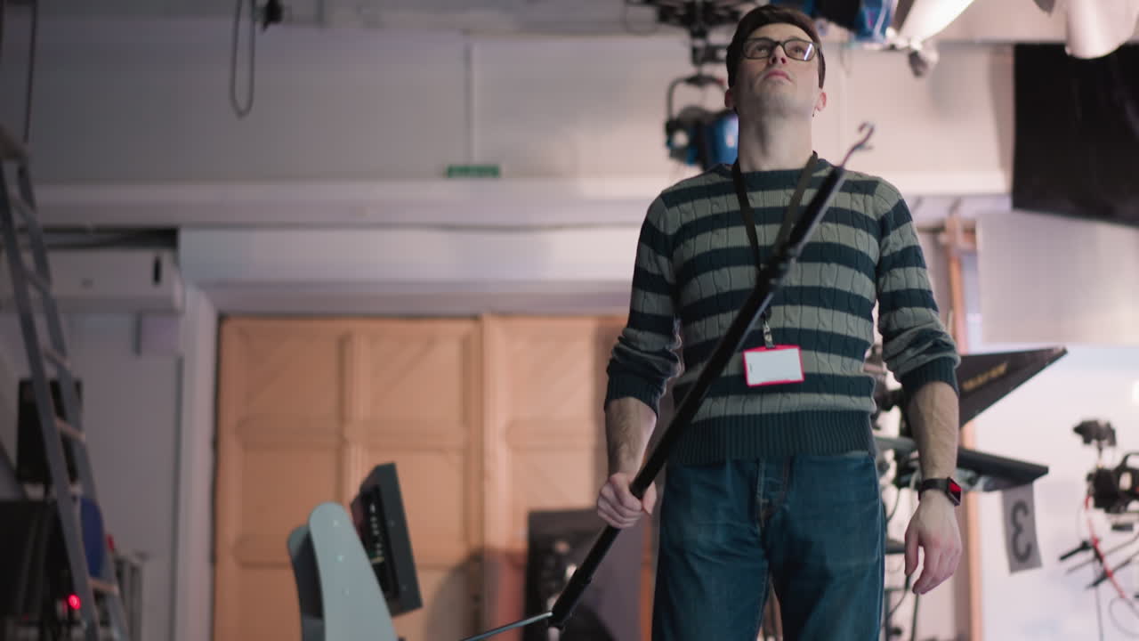 Technician adjusting equipment with boom pole in broadcast studio. Focused on lighting and camera setup while performing technical work in studio environment for filming production