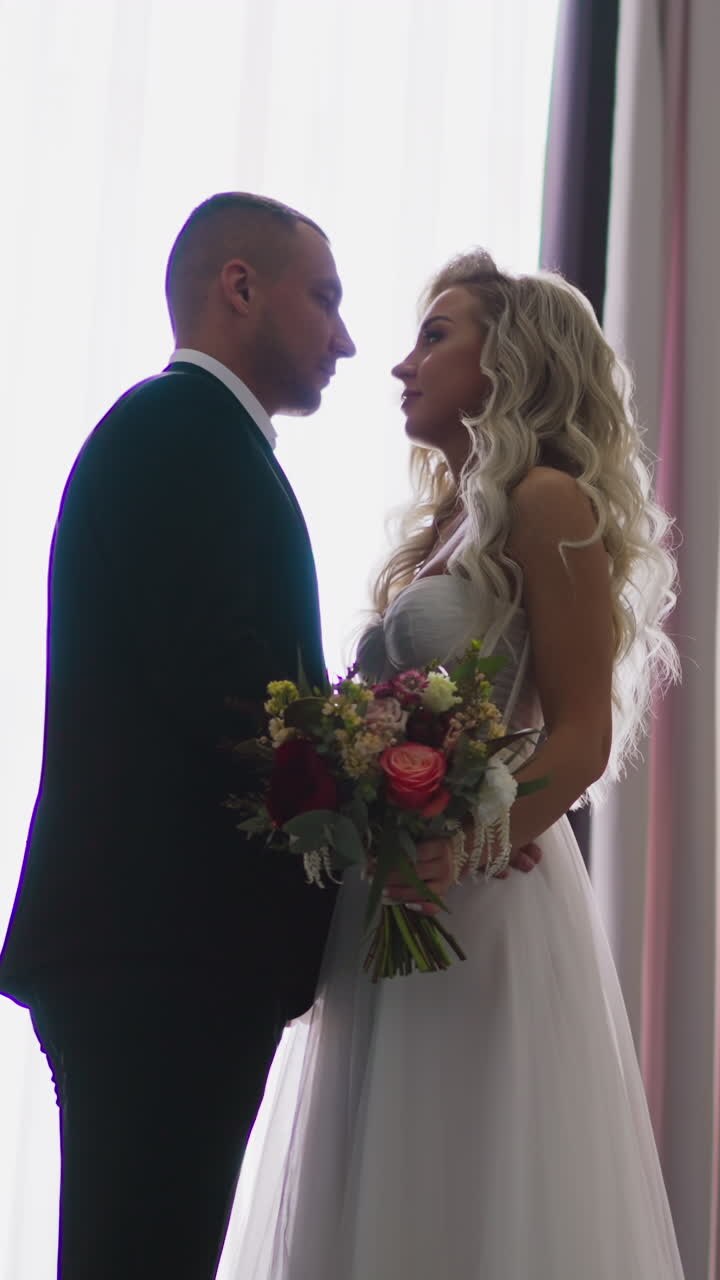 Loving pretty long haired bride with bouquet and positive groom stand on vintage staircase by window before wedding ceremony slow motion