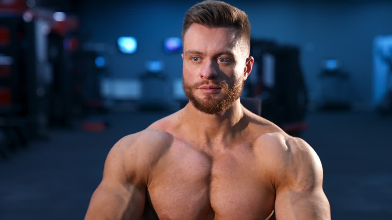 Portrait of bodybuilder indoors. Young bearded sportsman with athletic body sitting in gym. Muscular athlete without shirt. Front view.
