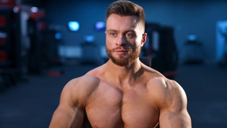 Portrait of bodybuilder indoors. Young bearded sportsman with athletic body sitting in gym. Muscular athlete without shirt. Front view.