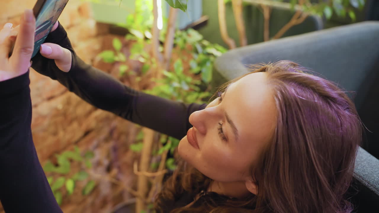 Lady with brown hair smiling while holding phone up to take picture surrounded by cozy indoor greenery and soft seating, with warm natural lighting and relaxed atmosphere in modern space