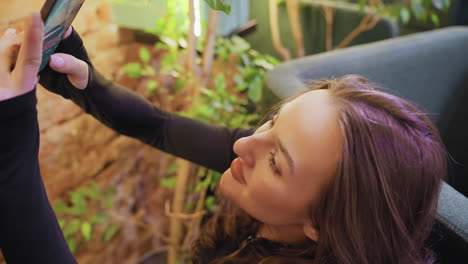 lady with brown hair smiling while holding phone up to take picture surrounded by cozy indoor greenery and soft seating, with warm natural lighting and relaxed atmosphere in modern space