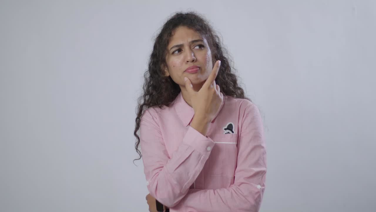 A Curly-haired South Asian woman in a pink shirt thoughtfully touches her face, lost in deep contemplation