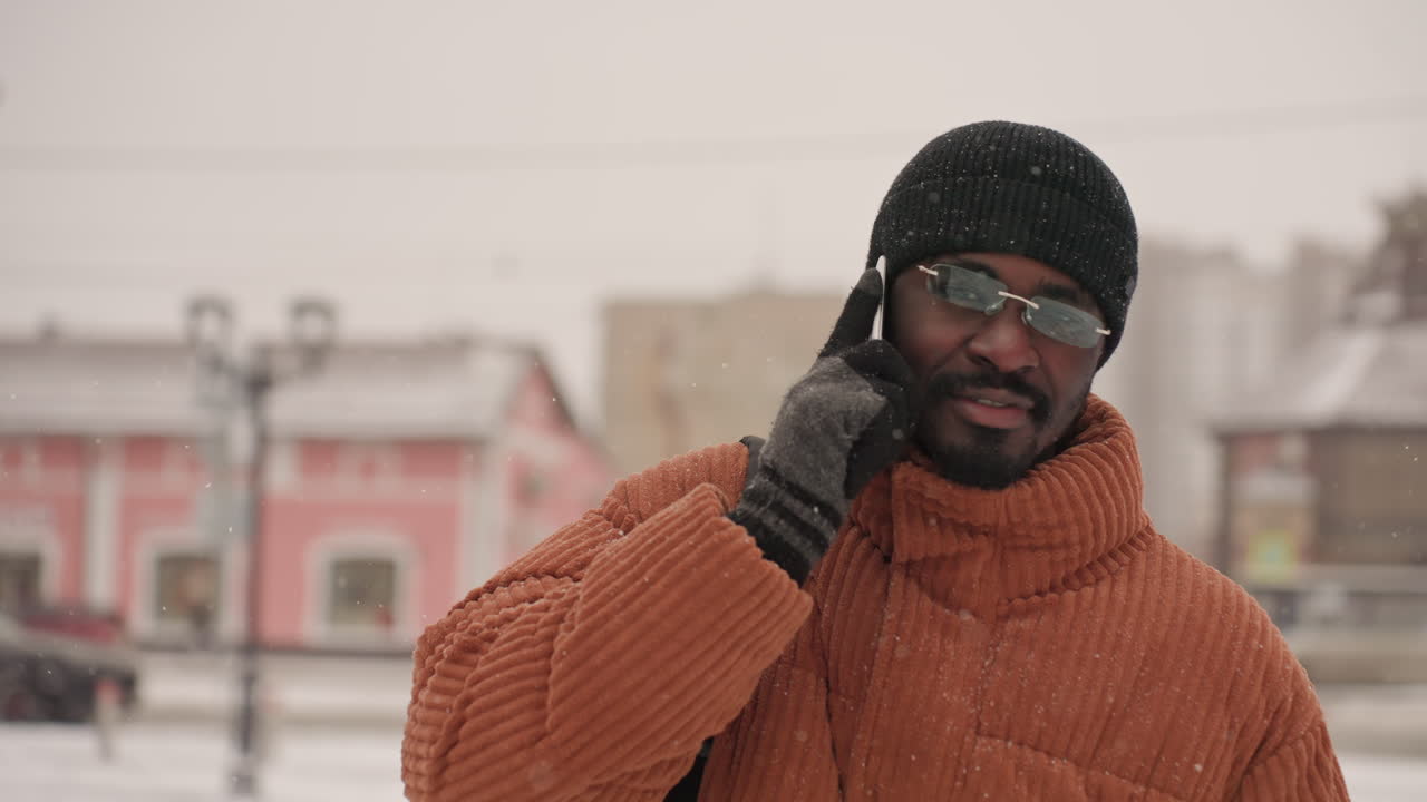 Uomo al telefono in una strada innevata che indossa un piumino arancione, un berretto nero e guanti, sorride e parla mentre cade una leggera neve, edifici urbani e lampioni sullo sfondo, ritratto casuale diurno